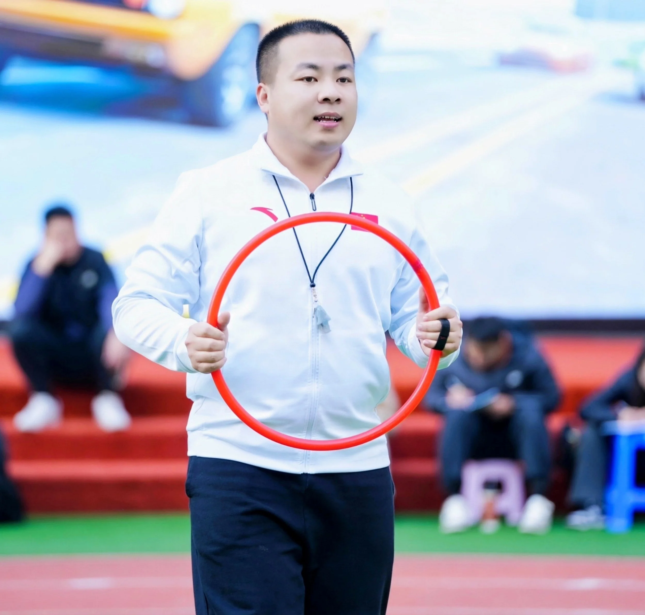 A young man holding a red hula hoop during an outdoor event, with other people sitting in the background.
