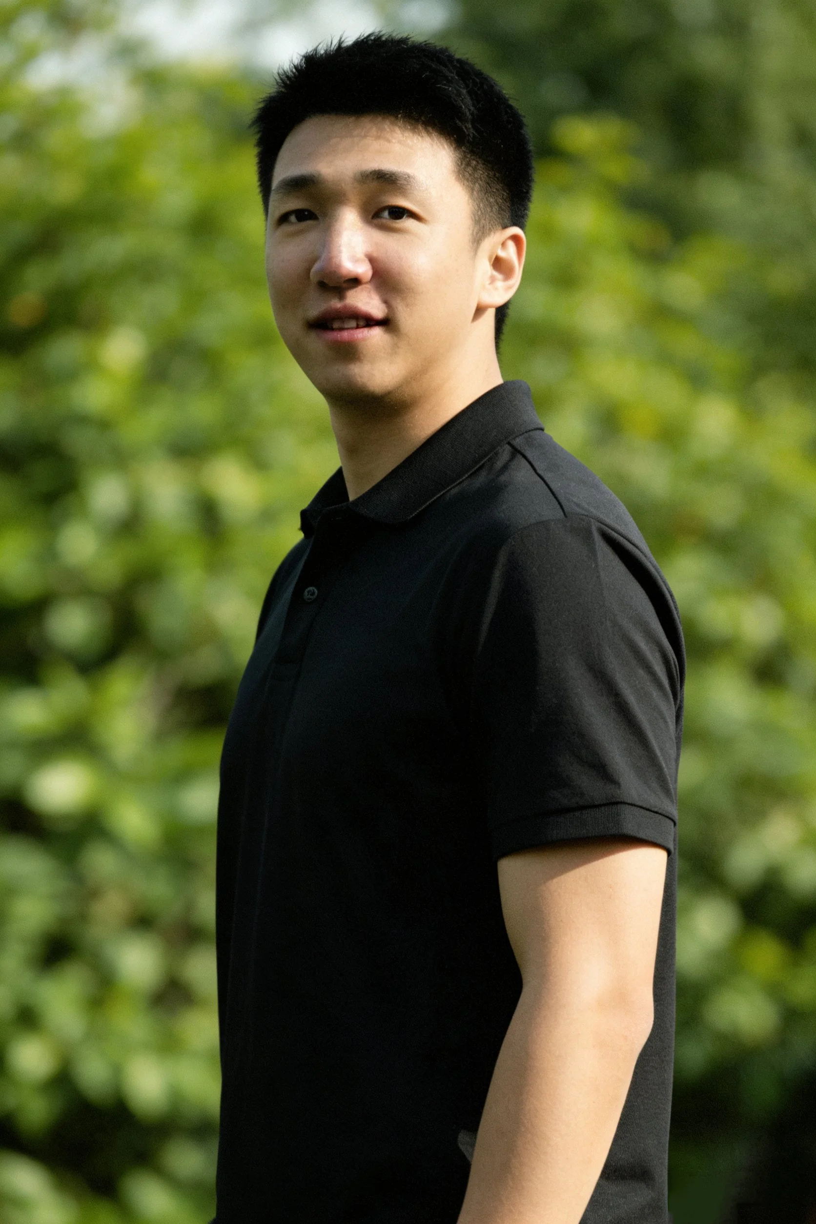 A young man with short black hair wearing a black polo shirt stands outdoors with green foliage in the background.