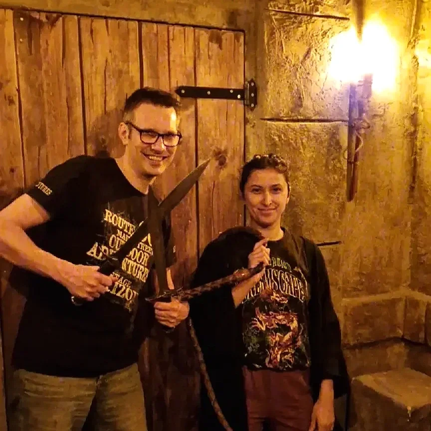 Two people, a man and a woman, are standing indoors against a rustic wooden and stone wall. The man on the left is smiling, wearing glasses and a black T-shirt with yellow text, and holding a sword. The woman on the right is smiling, dressed in a black jacket, a graphic T-shirt featuring a dragon, and holding a snake. Warm lighting illuminates the scene.
