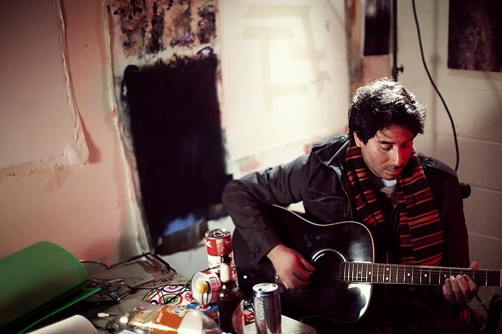 A man sitting at a table playing an acoustic guitar in a room with unfinished walls and various items on the table.