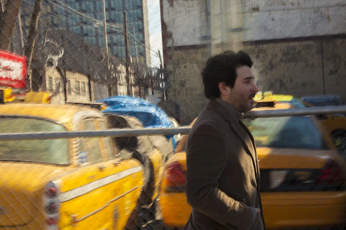 A man with dark hair and a beard walking past yellow taxis in an urban street scene.