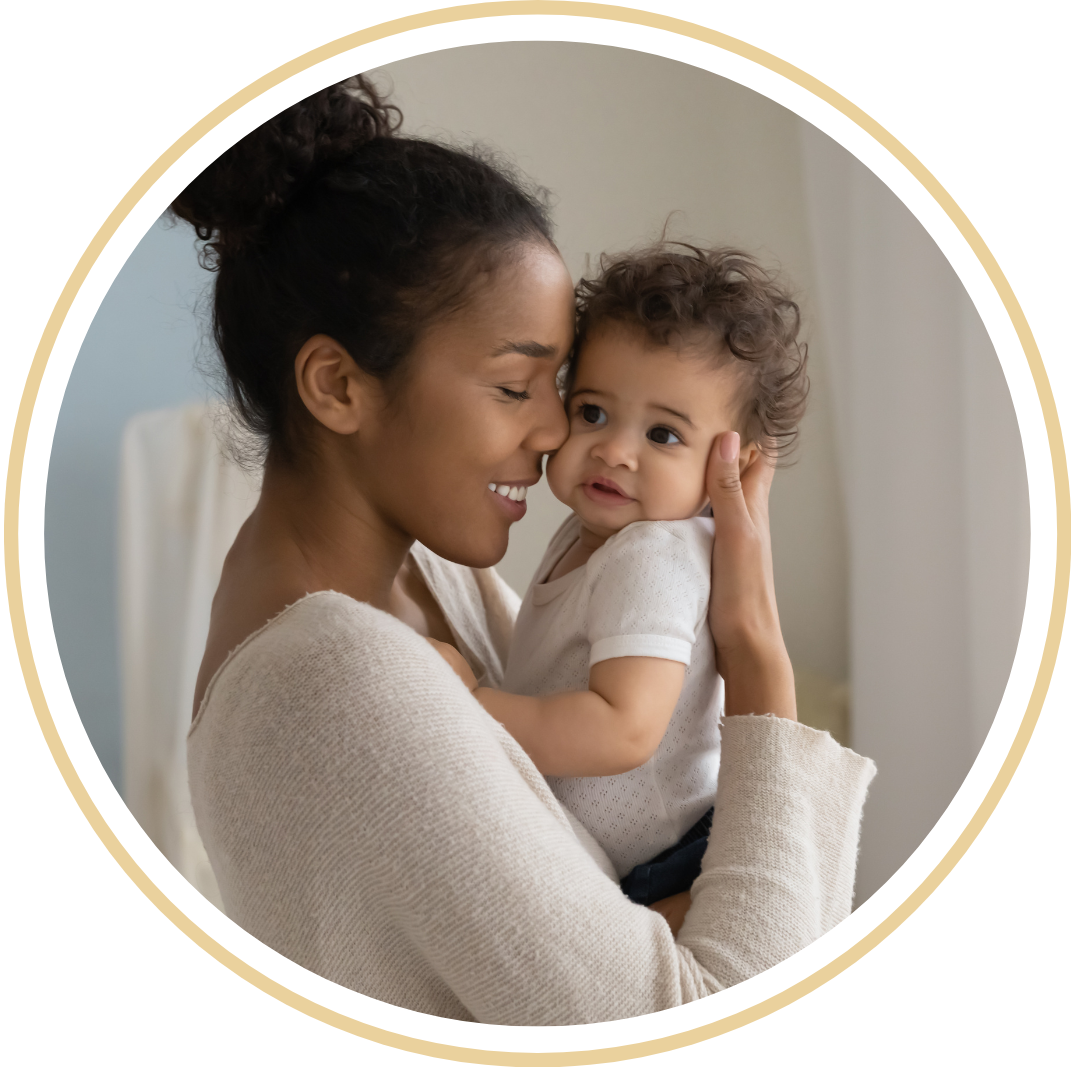 A soft-focus photograph of a mother holding her baby close in a warm, dimly lit room. She has a peaceful expression as she offers a gentle, soothing embrace, reflecting GJS’s connection-based approach to sleep support.