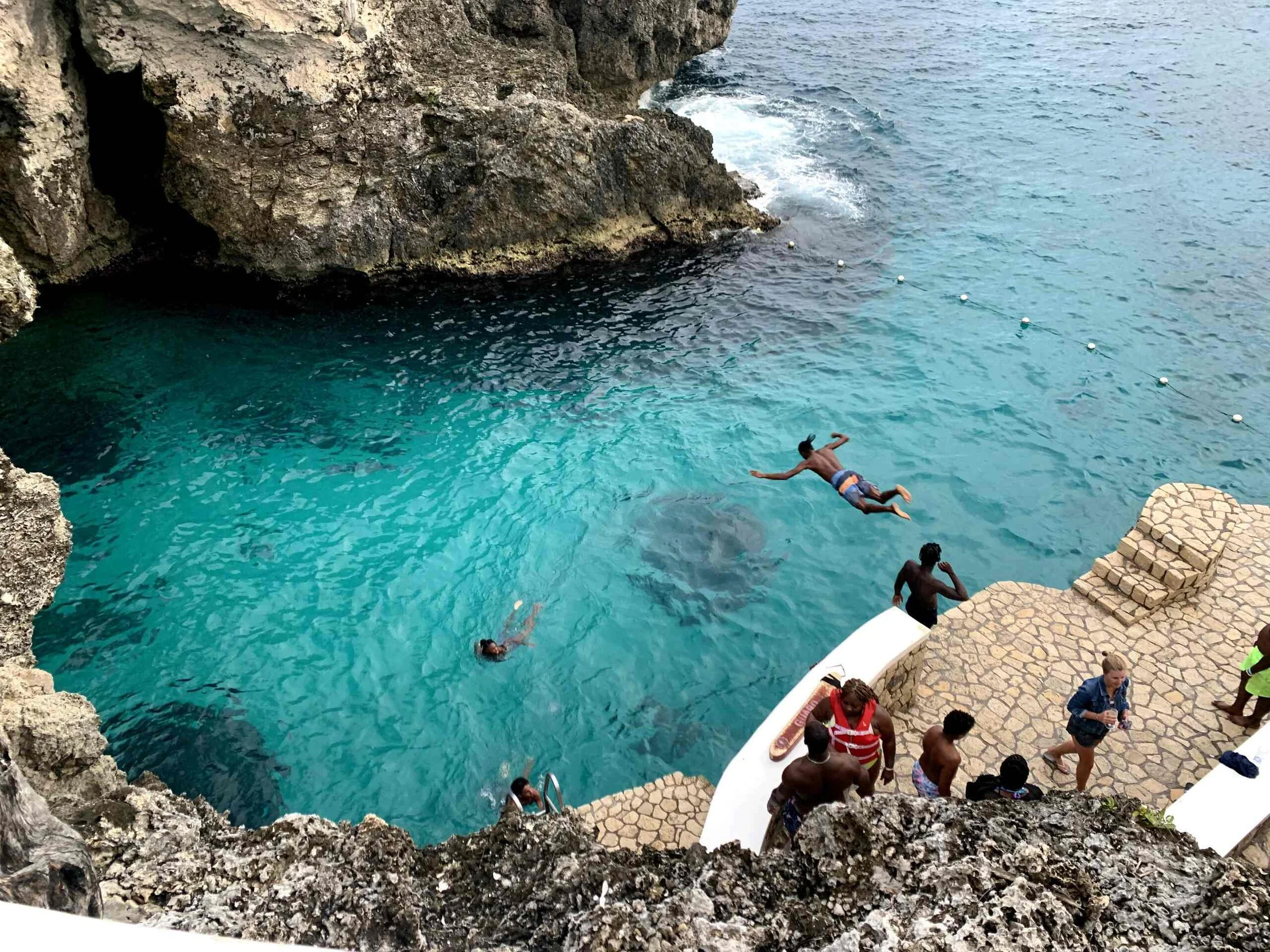 People swimming and jumping into a turquoise water at a rocky seaside cove with stone steps leading into the water and a few onlookers on the dock.