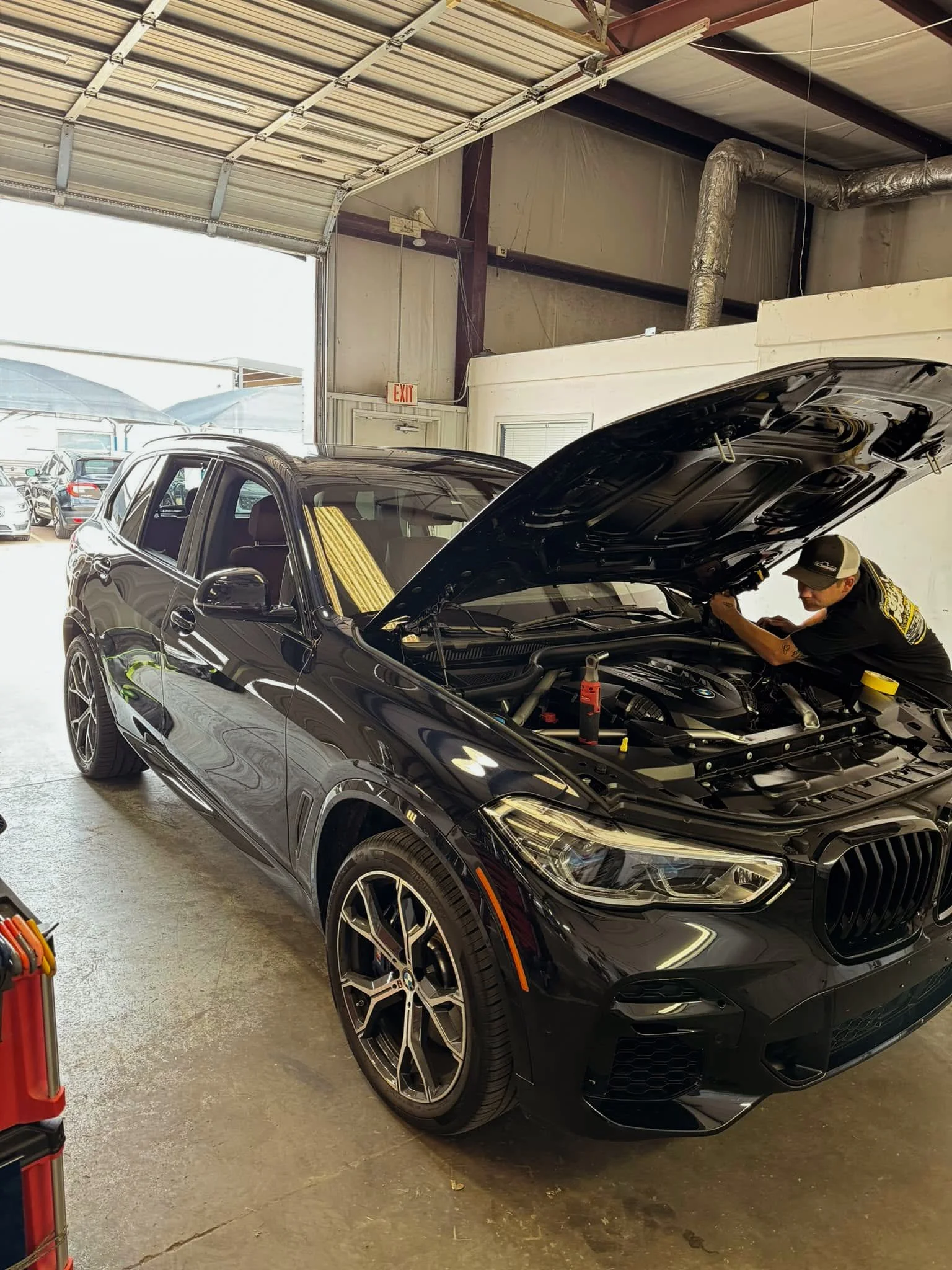 A black BMW SUV with mounted wheels, open hood, and a mechanic working on the engine inside a garage.