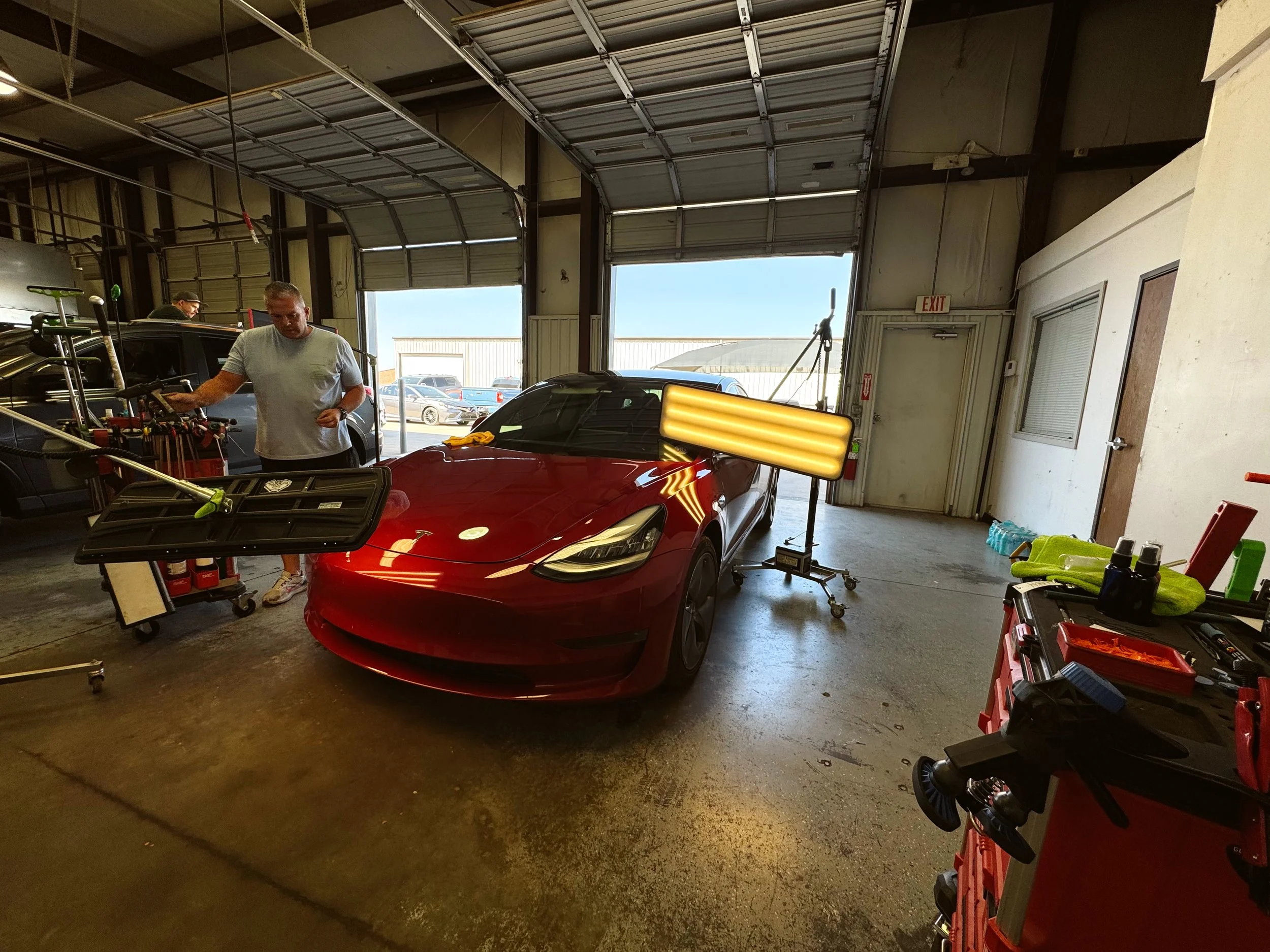 Inside a garage or workshop with a red Tesla car in the center, a man working on the vehicle, and various tools and equipment around, including a large yellow light on a stand close to the car.