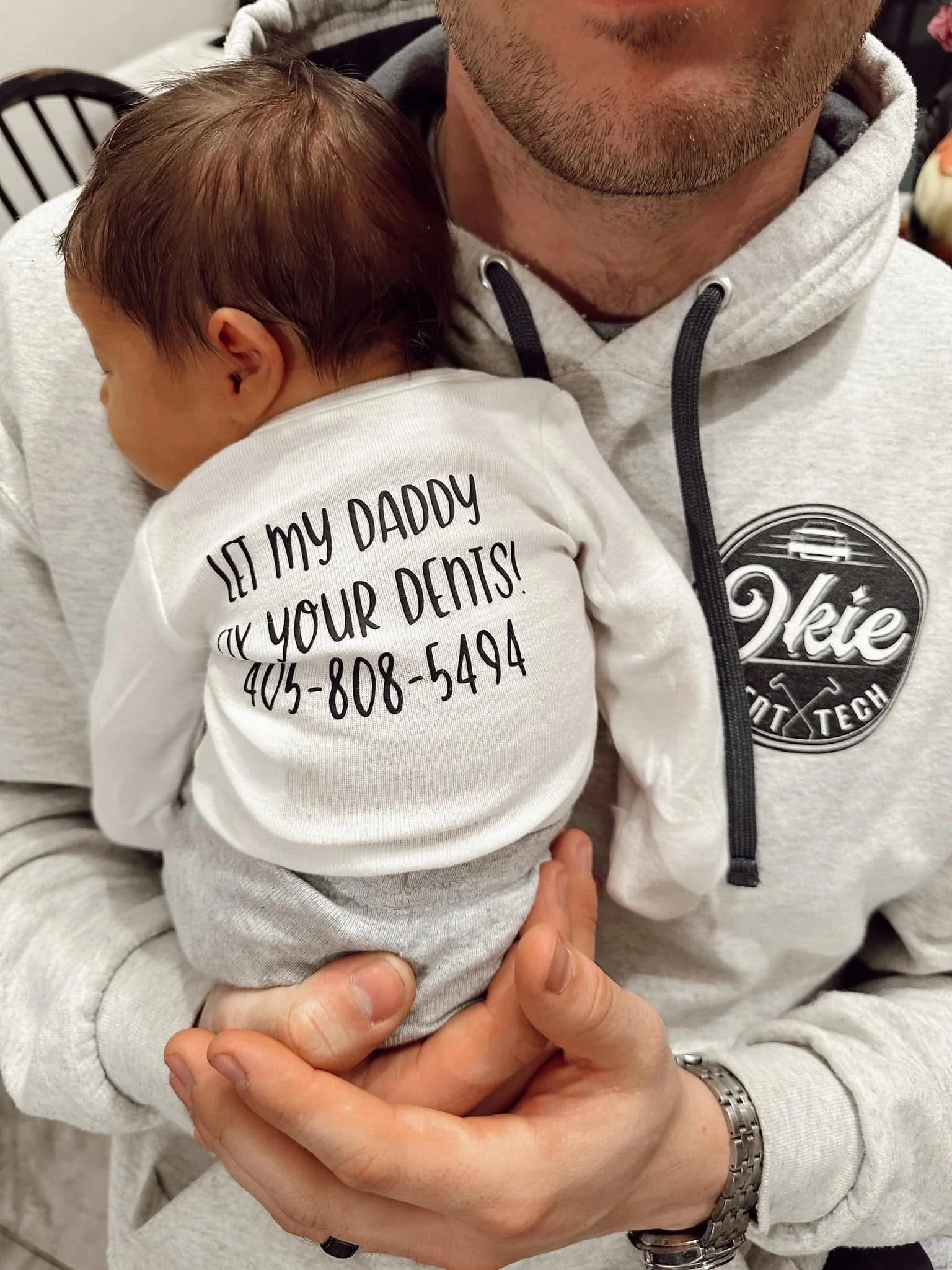 A man holding a sleeping baby on his chest, wearing a white shirt with text and a phone number printed on the back. The man is wearing a gray hoodie with a logo that says '2kie' and 'Tech'. The baby is dressed in gray pants and has brown hair.