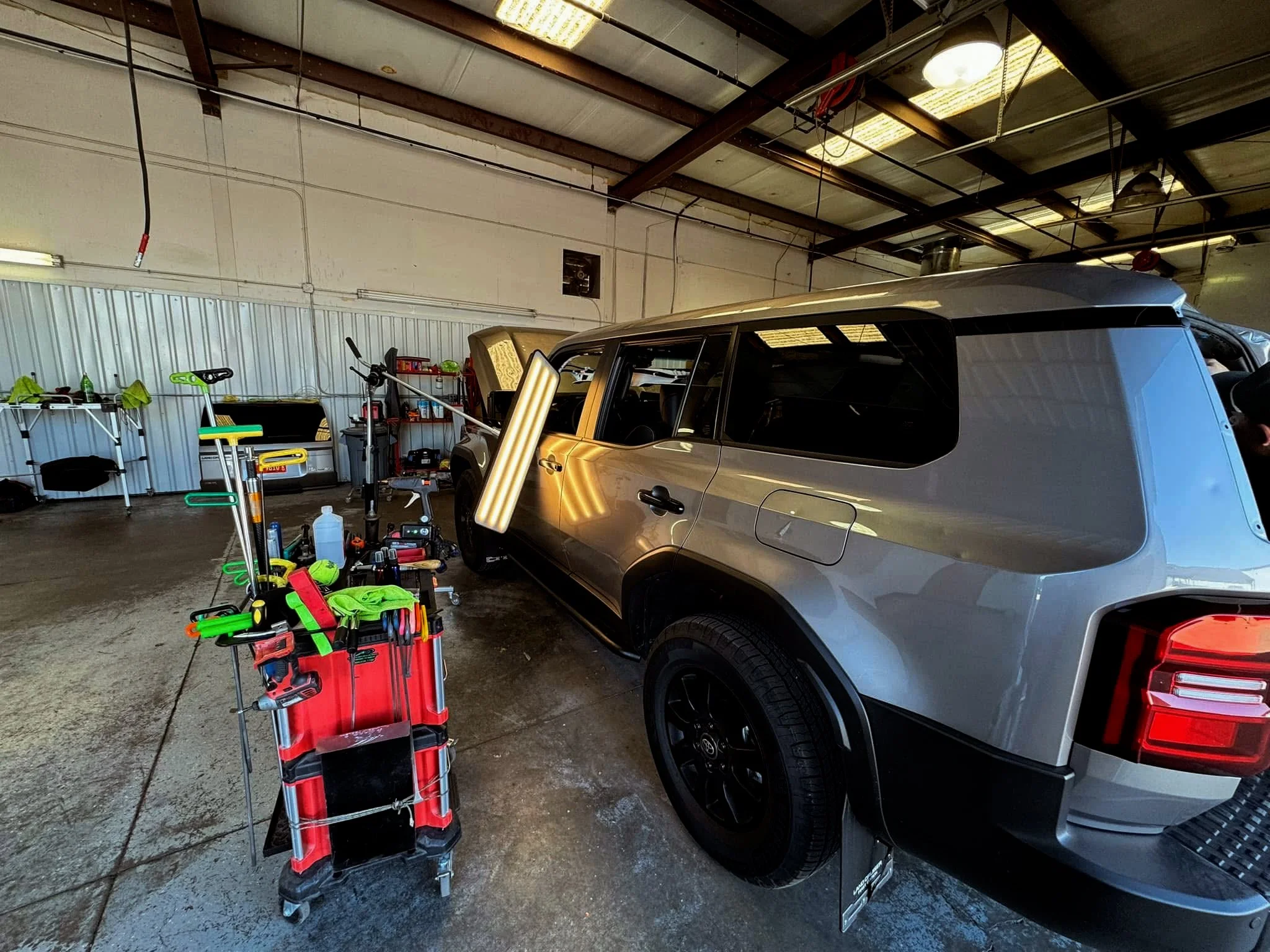 Inside a garage with a silver SUV parked on the right, surrounded by a variety of car detailing tools and equipment on a rolling cart and against the wall.