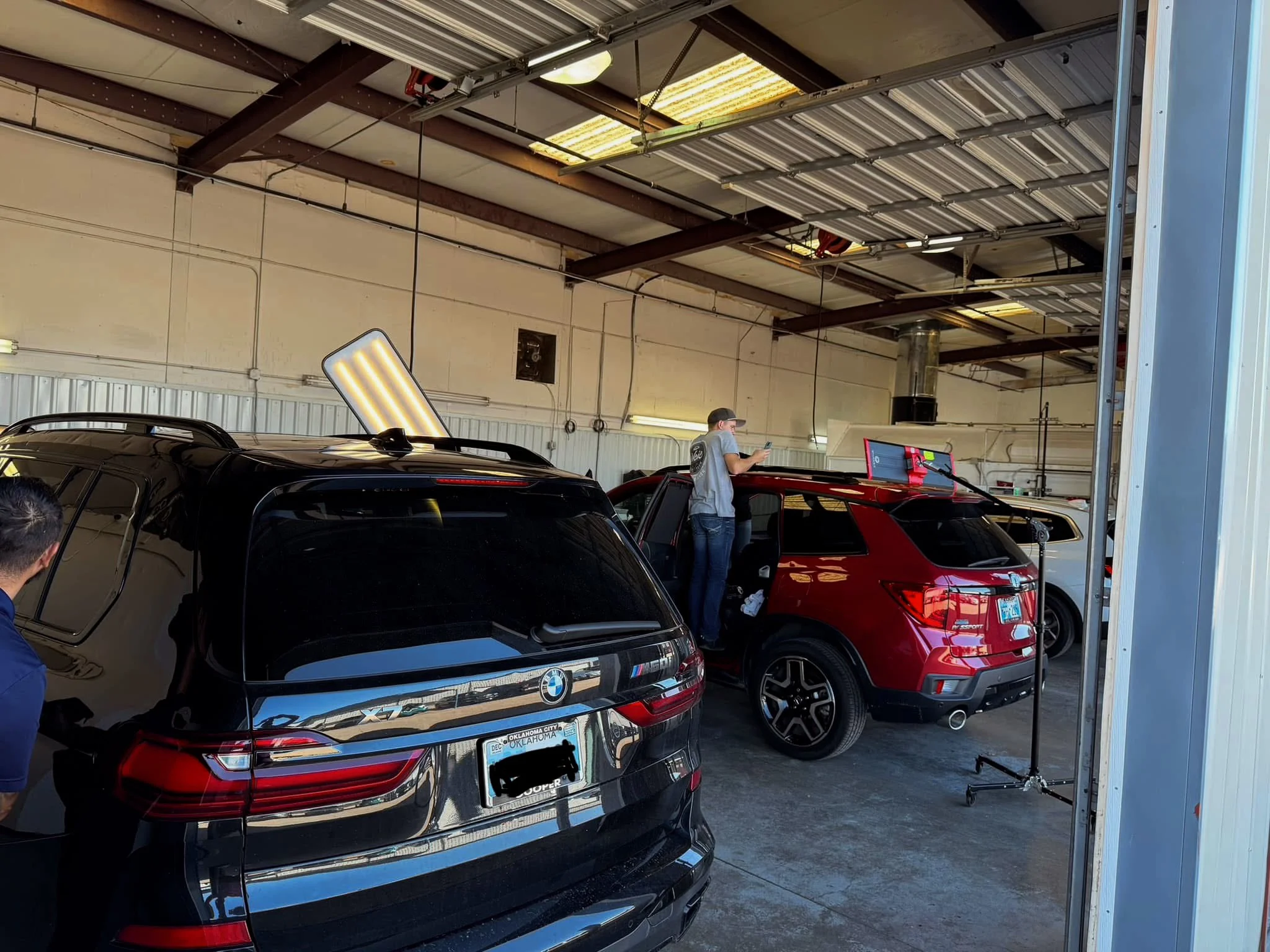 Inside a garage or auto shop with a black BMW SUV and a red vehicle being worked on. Two people are present, one inspecting the red vehicle and another partially visible near the BMW. Equipment and lighting are visible on the ceiling.