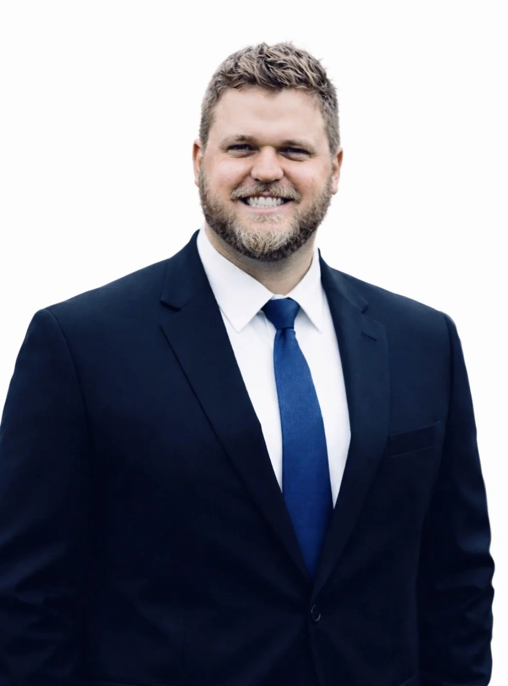 A man with a beard and curly hair smiling, wearing a black suit, white shirt, and blue tie, posing against a plain white background.