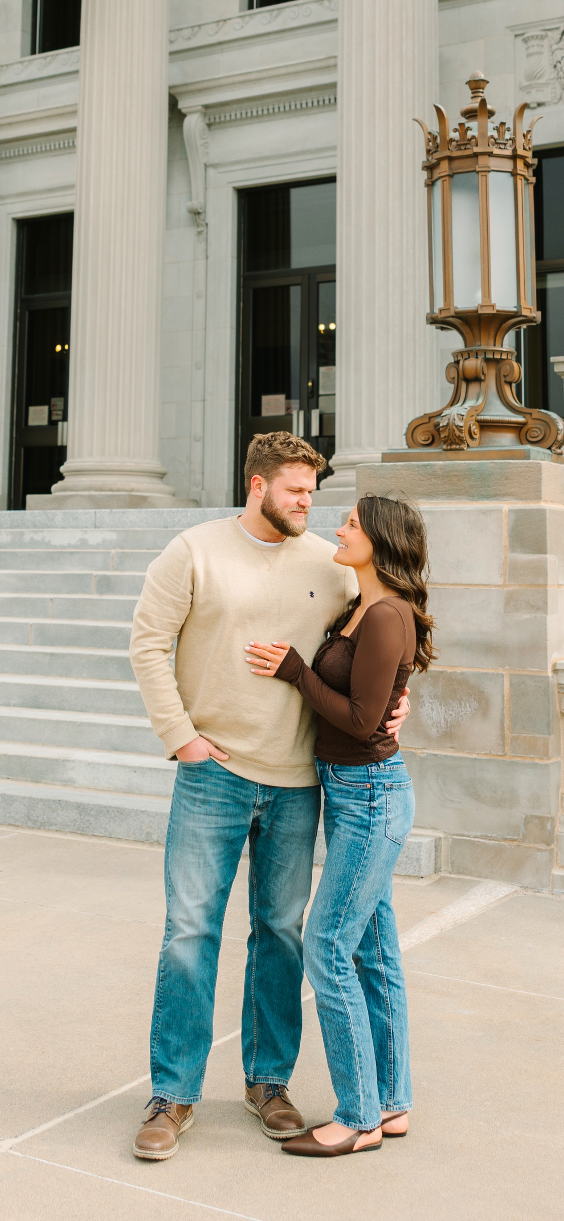 A man and woman standing close together on a sidewalk in front of a large, classical-style building with columns. They are gazing at each other affectionately. The man is wearing a beige sweater and blue jeans, and the woman is wearing a dark brown long-sleeve top and blue jeans.