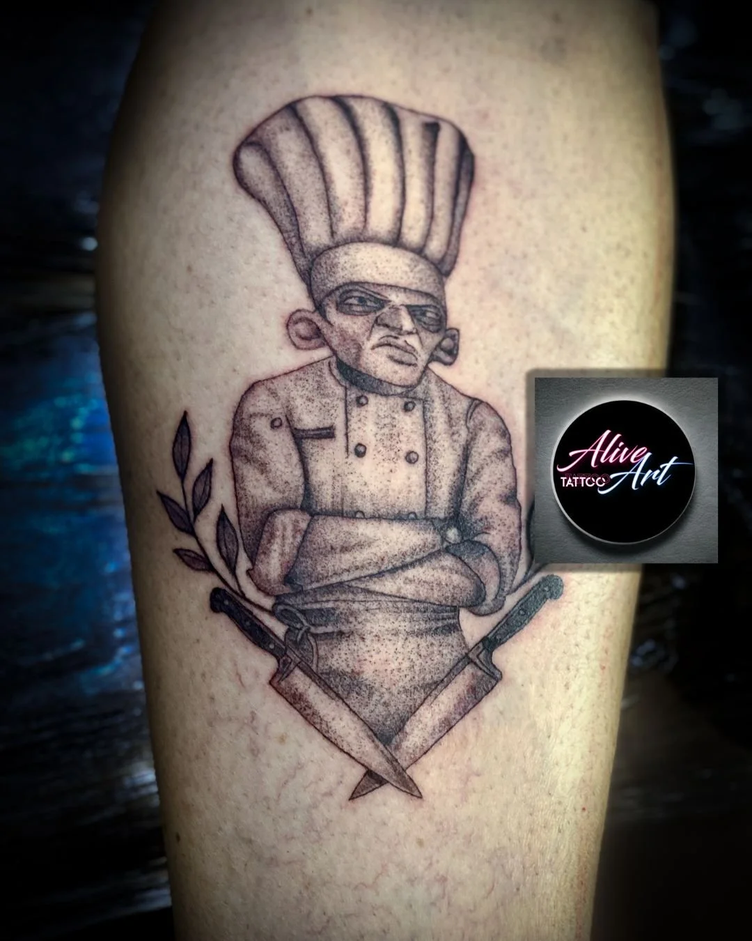 Tattoo of a stern chef with crossed arms, wearing sunglasses and a tall chef hat, with two knives crossed below, and a small branch with leaves to the side.