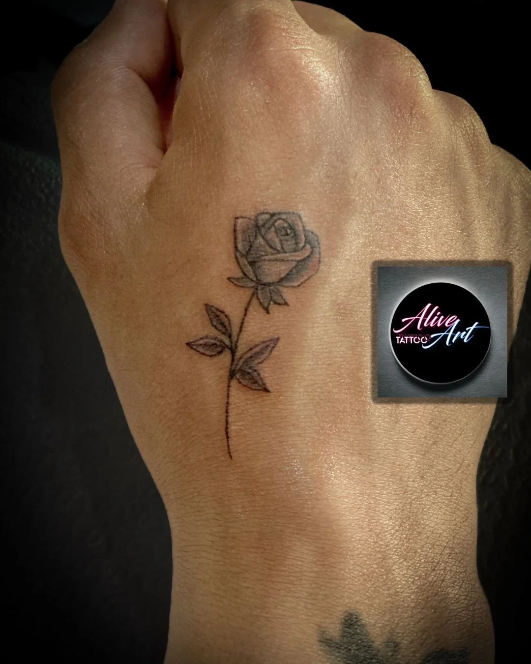 A black and grey tattoo of a rose on the back of a person's hand with the logo 'Alive Art TATTOO' in the corner.