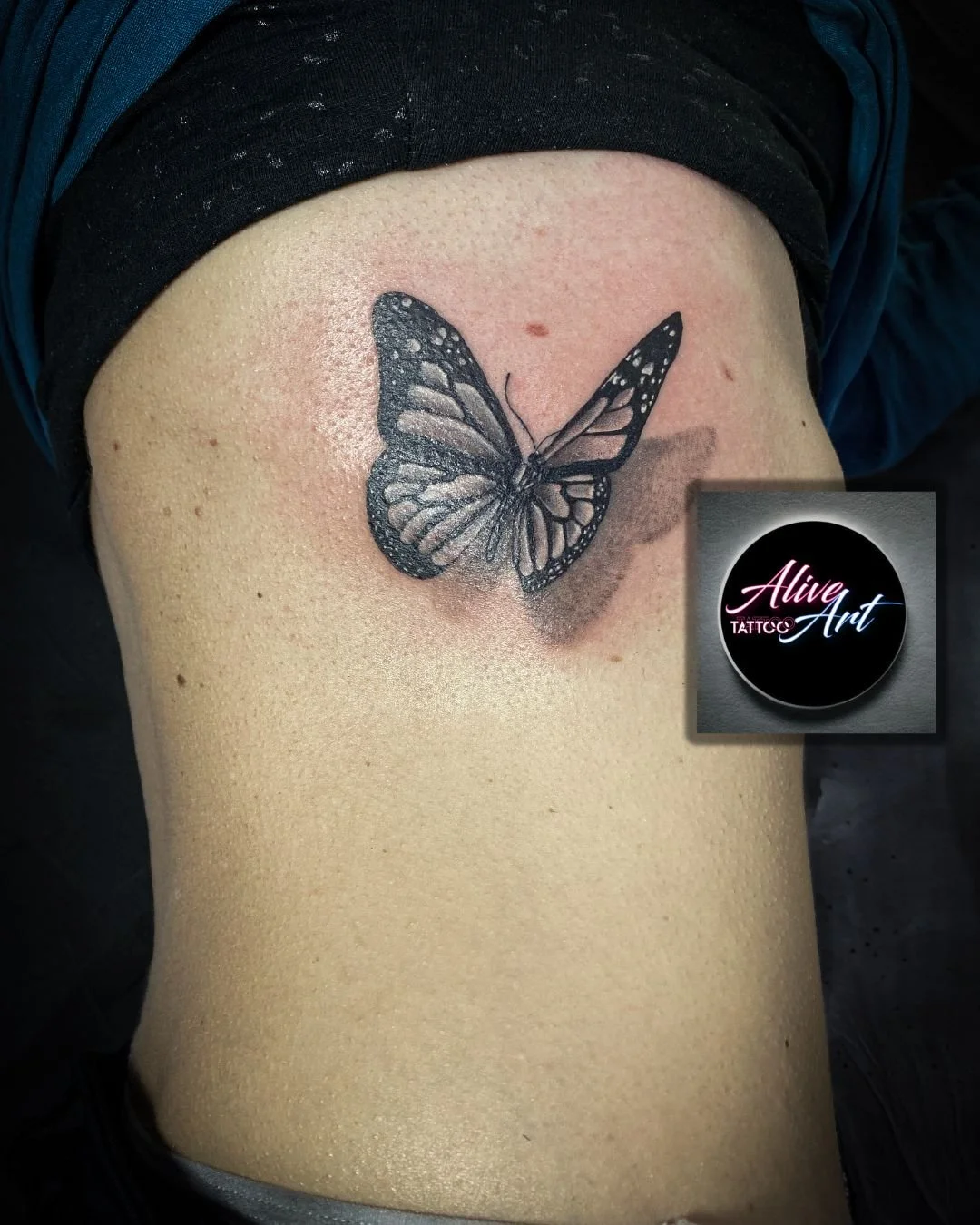 Black and gray butterfly tattoo on a person's side ribcage, with an 'Alive Art' tattoo studio logo in the corner.