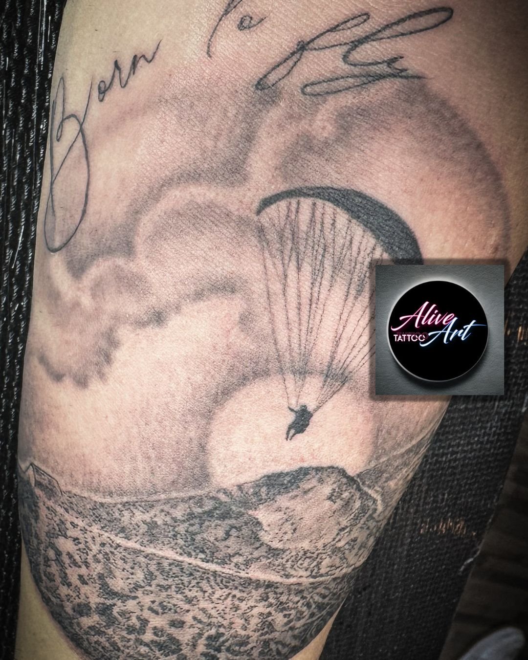 Black and gray tattoo of a person parachuting over a landscape with mountains and clouds, with a signature or writing above.