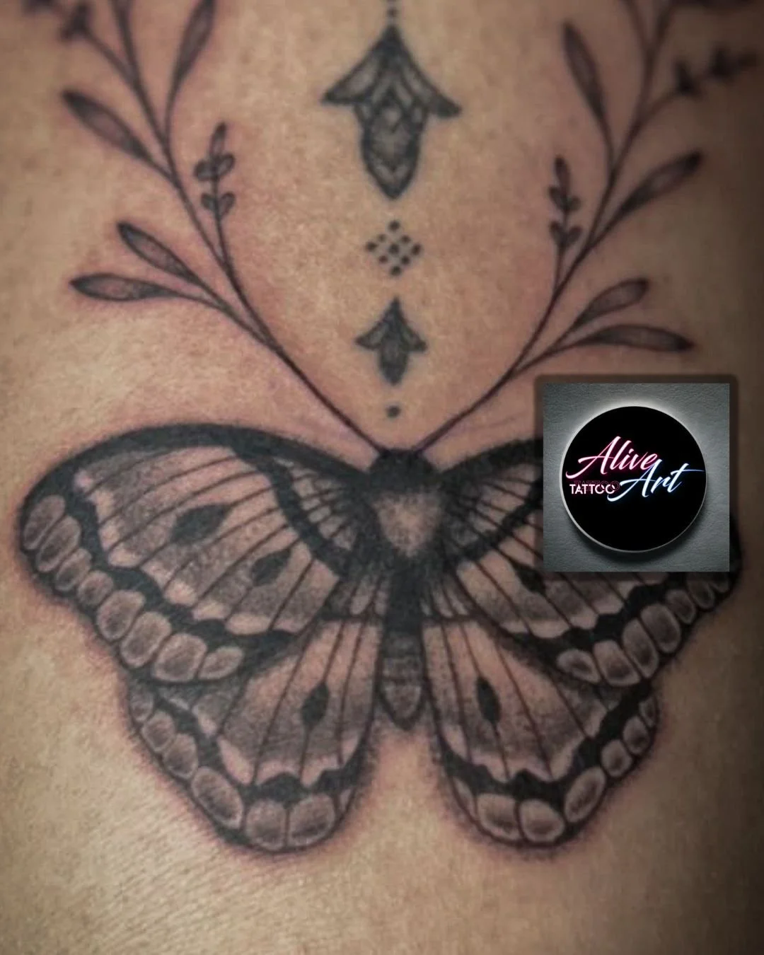 Tattoo of a butterfly with decorative geometric and floral elements above it, on skin.