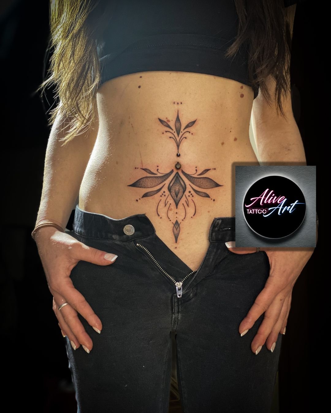 A woman with a tattoo on her lower abdomen featuring a symmetrical, floral design with black ink and dot accents, with the logo of Alive Art Tattoo in the bottom right corner.
