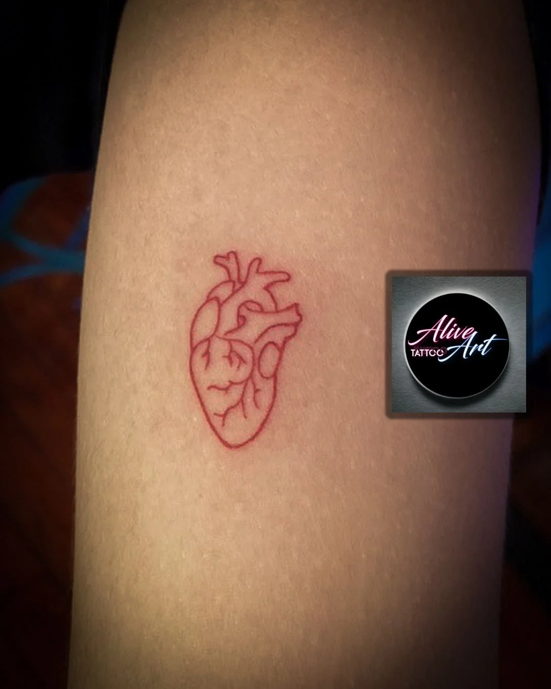 Red outline tattoo of a human heart on skin, with a small logo that says "Alive Art TATTOO."