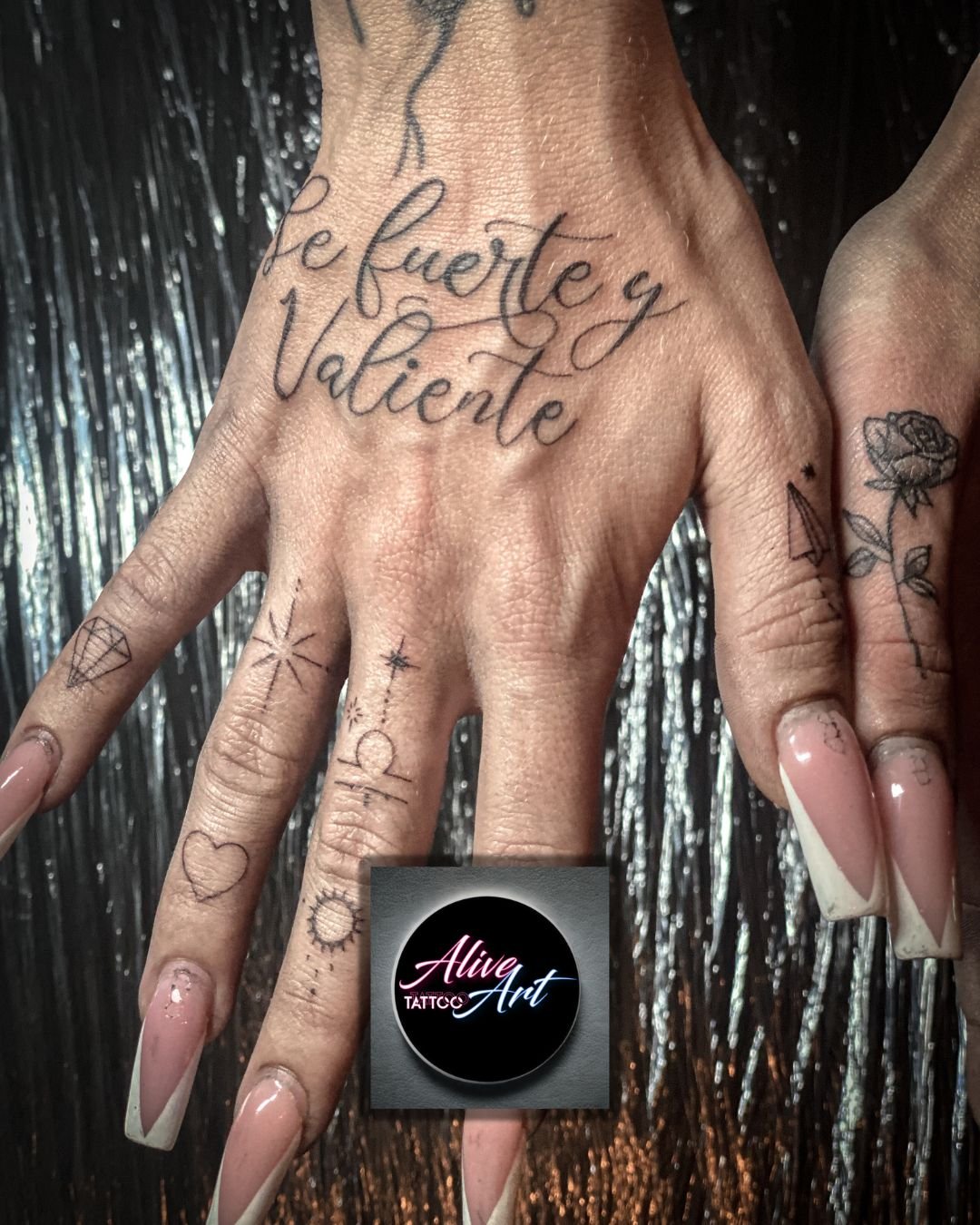 Close-up of a hand with multiple tattoos, including a quote in Spanish, a rose, geometric designs, and a heart, against a black shiny background, with long manicured nails.