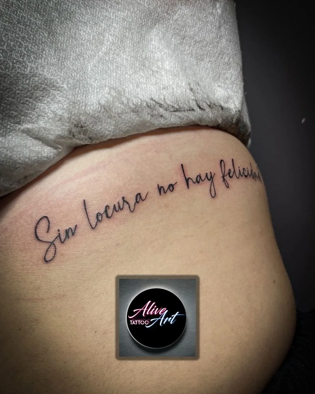 Close-up of a tattoo on a person's side with the inscription in Spanish, 'Sin locura no hay felicidad,' written in cursive. Part of the person's clothing and a circular sticker with the text 'Alive Art TATTOO' are also visible.