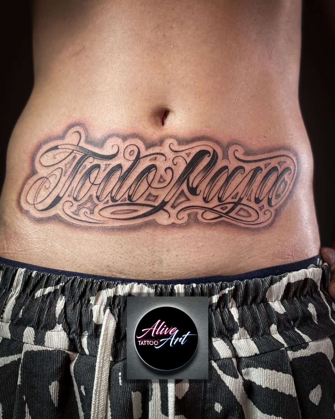 Tattoo on person's lower abdomen with cursive writing that says 'Stay Ready', above a pair of camouflage shorts and a black button with 'Alive Art' logo.