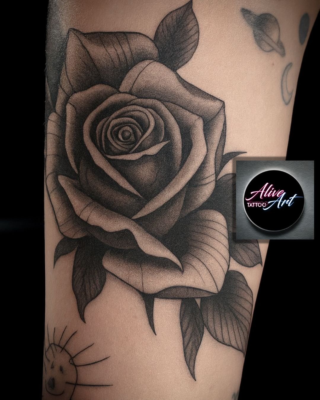 Black and gray tattoo of a detailed rose with leaves on skin, with smaller tattoos of a smiling sun and other designs nearby.