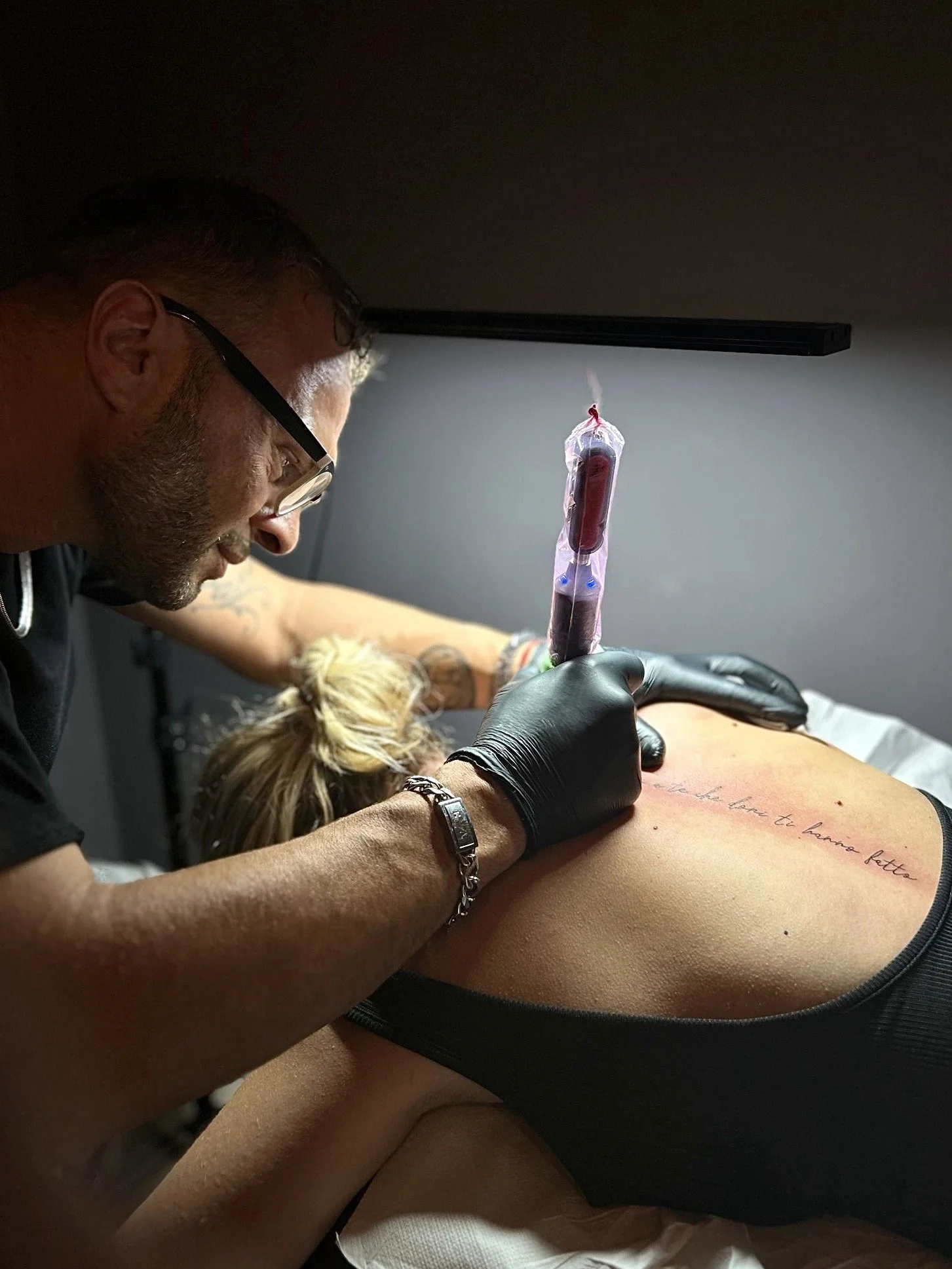 Tattoo artist in black gloves tattooing script on a woman's back in a tattoo studio.