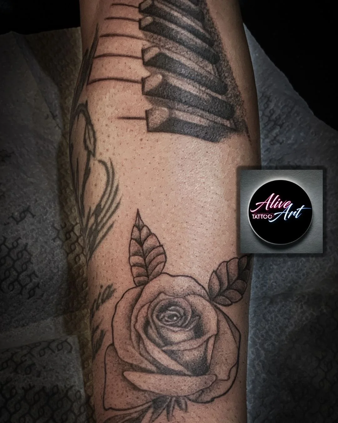 Tattoo of a rose with leaves on a person's forearm, alongside other tattoo designs including piano keys and a feather. The tattoo is black and gray and detailed.