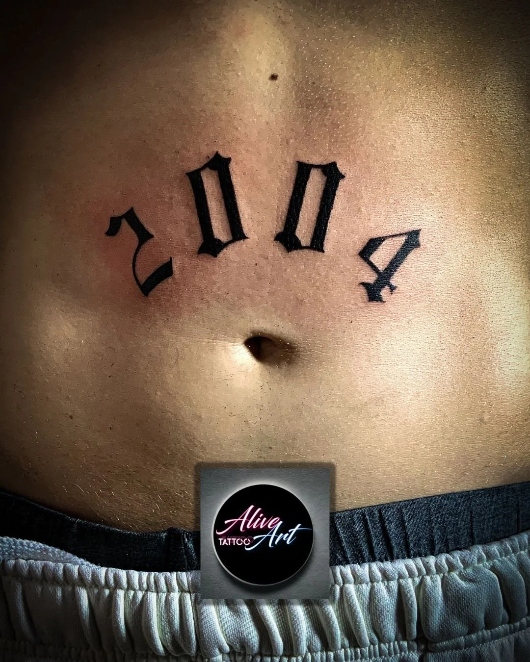 A tattoo of Roman numerals on a person's stomach, above the navel, with the words 'Alive Art' below the tattoo.