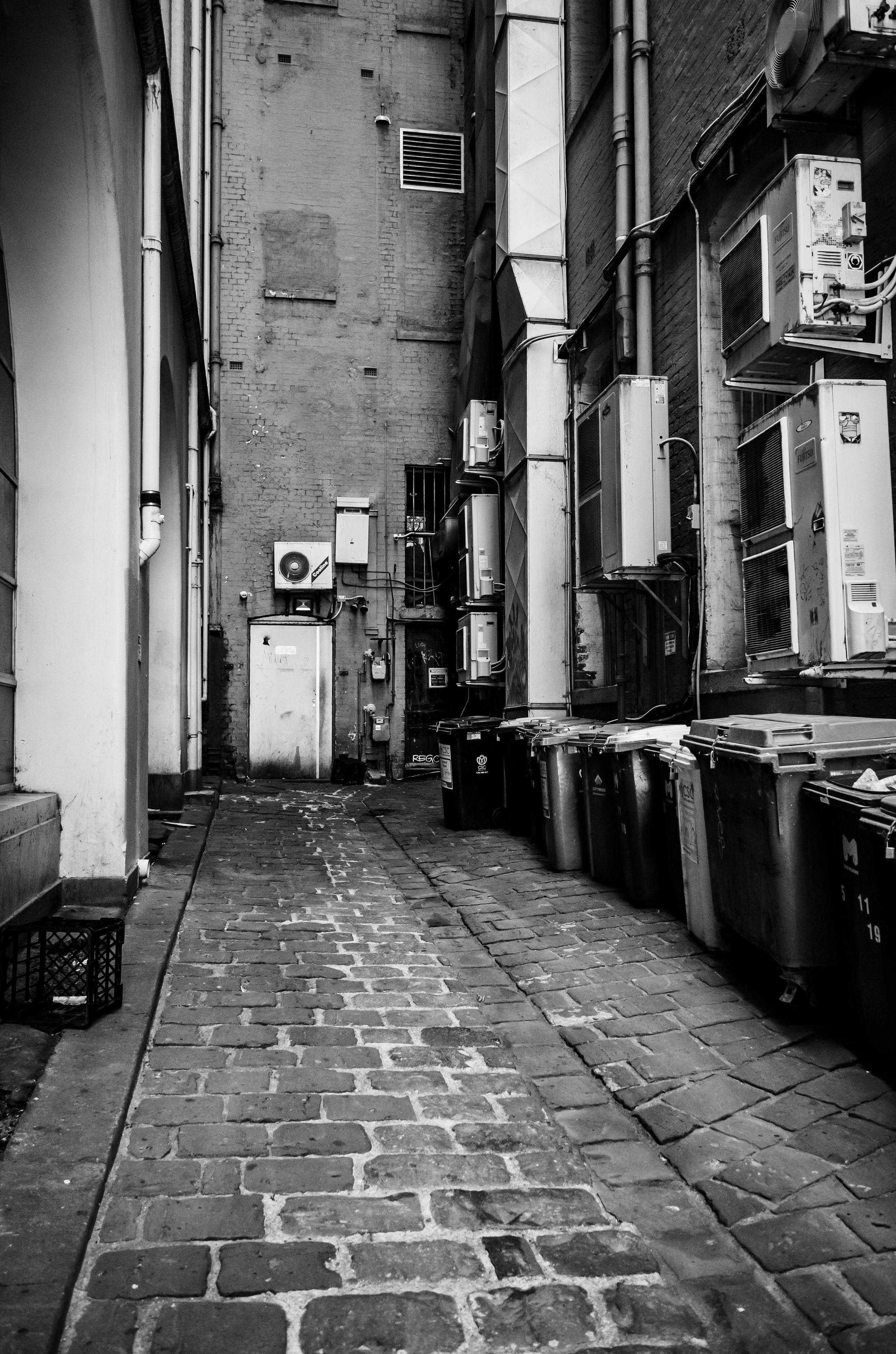 Black and white photograph of a narrow alleyway with cobblestone pavement, brick walls, and multiple air conditioning units mounted on the wall on the right side, with several trash bins lined up along the right edge.