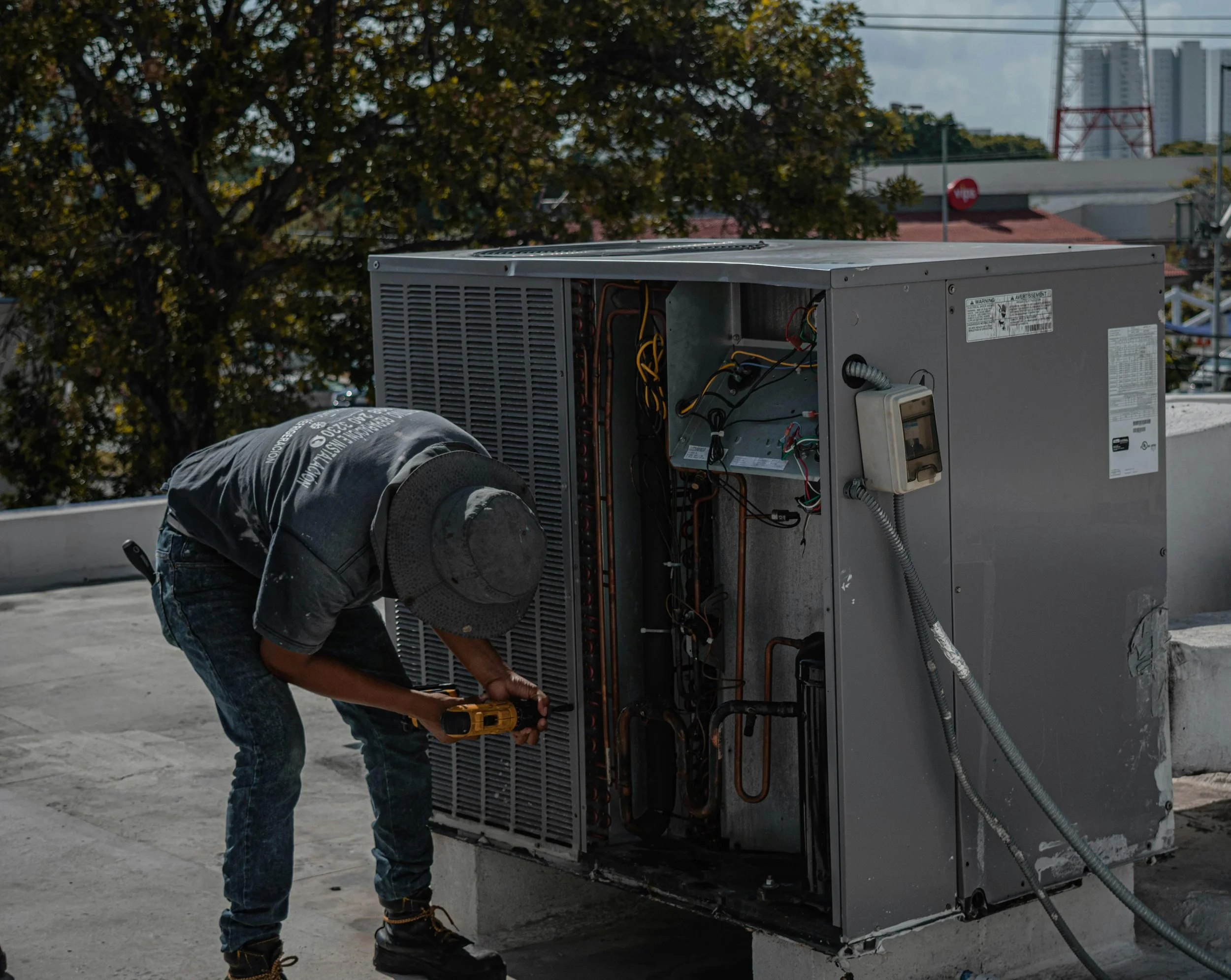 A worker in a hat and jeans is repairing or maintaining an outdoor HVAC unit on a rooftop, with trees and city buildings in the background.