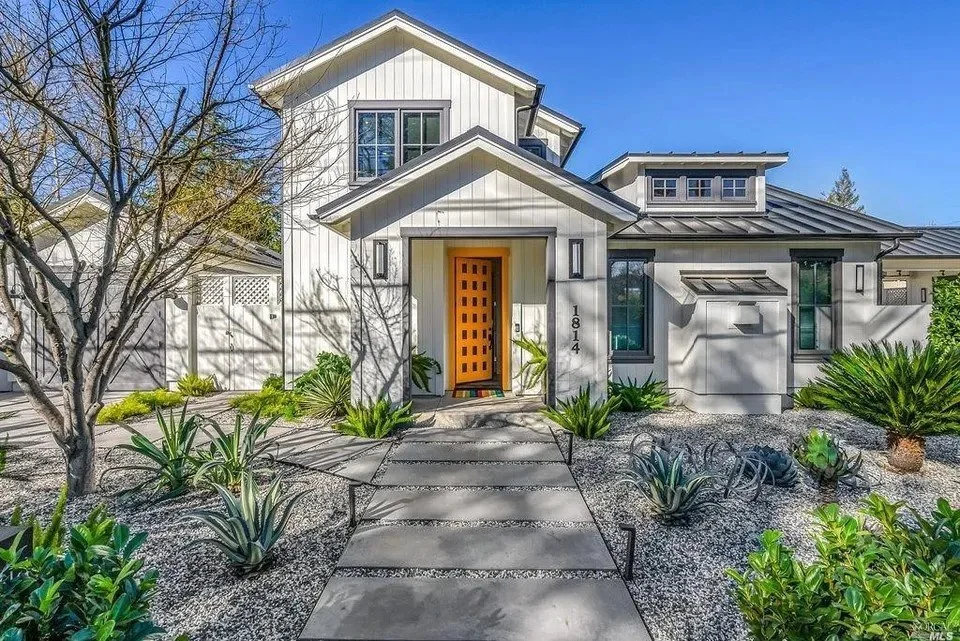 Modern white house with a wooden front door, surrounded by desert landscaping including aloe plants, a small tree, and shrubs, under a clear blue sky.