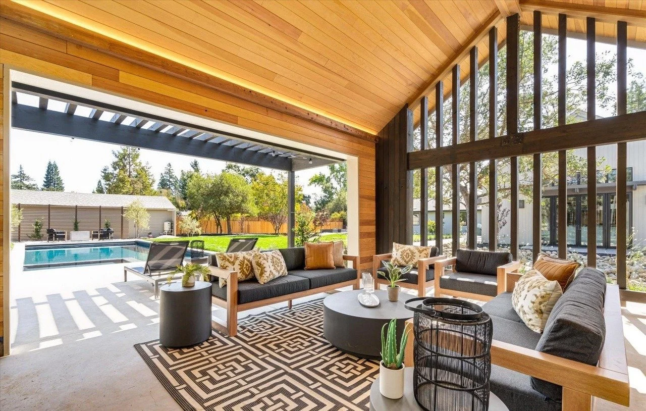 Indoor-outdoor living space with wooden ceiling, large windows, and patio furniture, overlooking a backyard swimming pool.