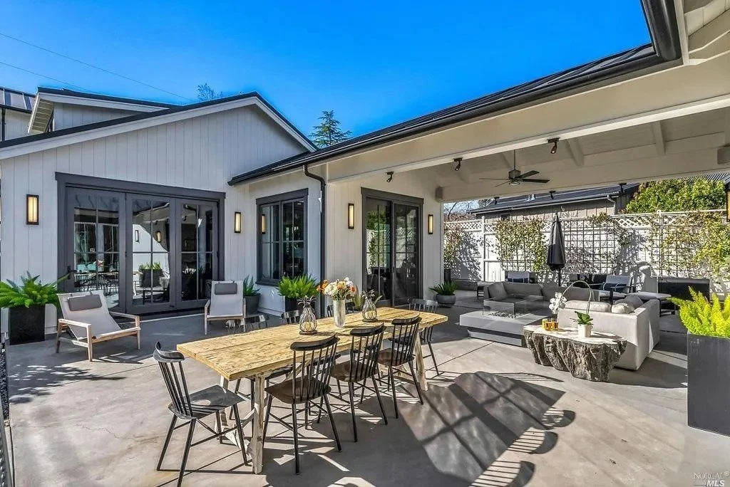 Outdoor patio with a wooden dining table, black chairs, and a seating area with gray sofas, potted plants, and an umbrella under a covered roof