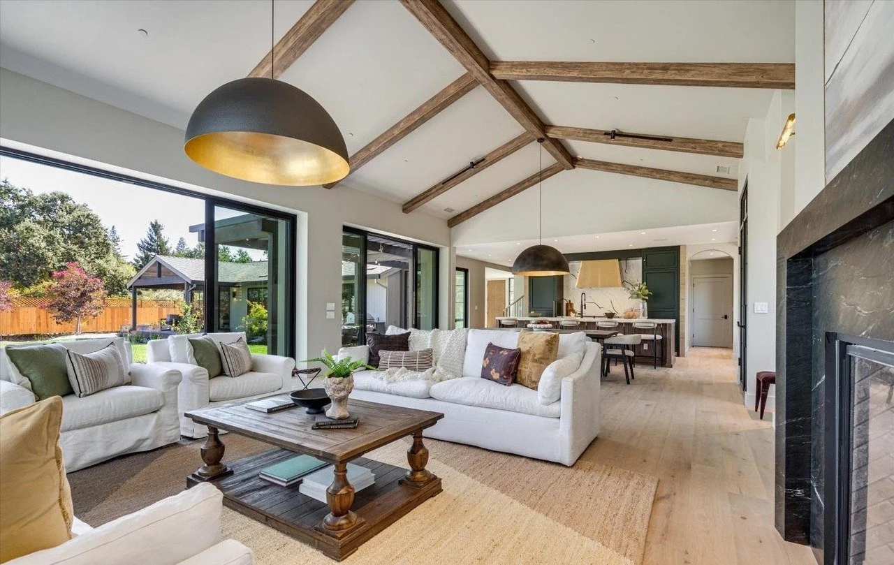 Open-concept living room and kitchen with vaulted ceilings, exposed wooden beams, large windows, two black pendant lights, white sofas with various pillows, wooden coffee table, black fireplace, and sliding glass doors leading to a backyard with trees and a wooden fence.