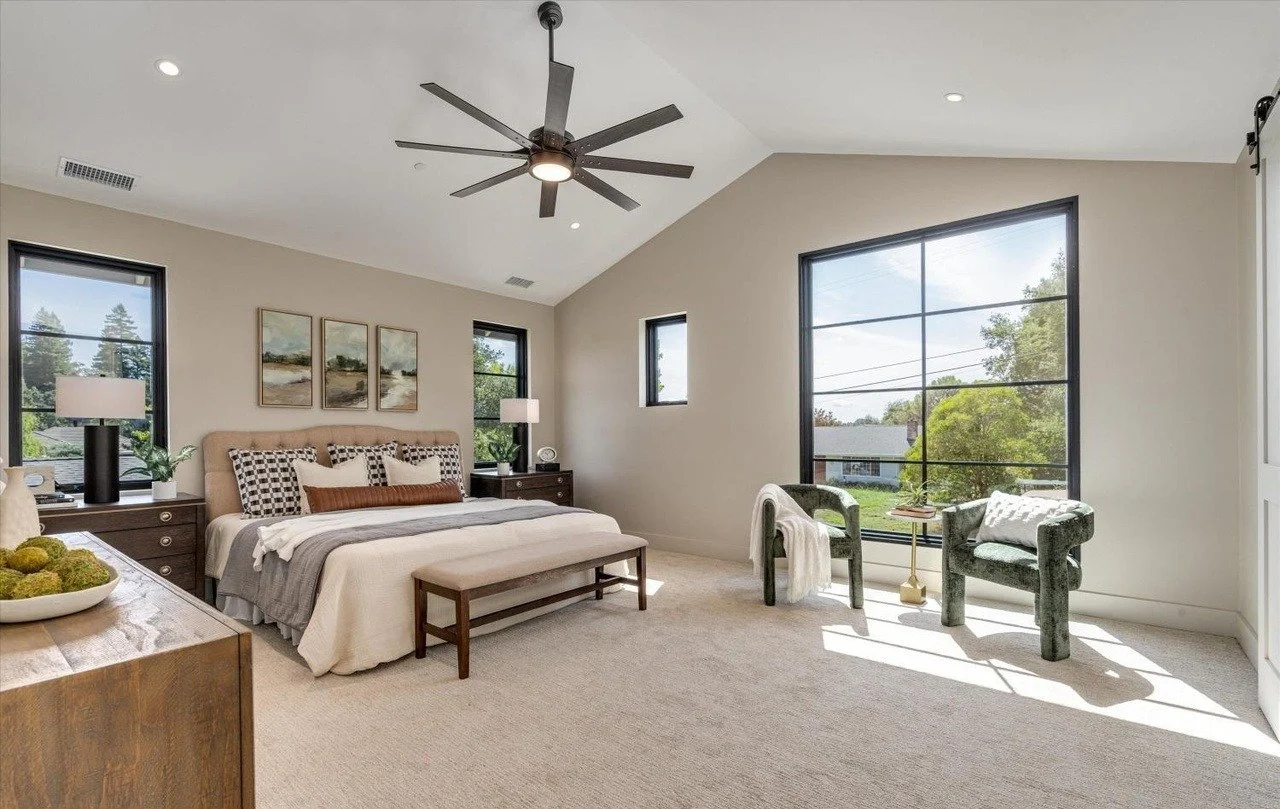 Spacious bedroom with large window, ceiling fan, bed with multiple pillows, two bedside tables with lamps, a bench at the foot of the bed, and two green armchairs near the window.
