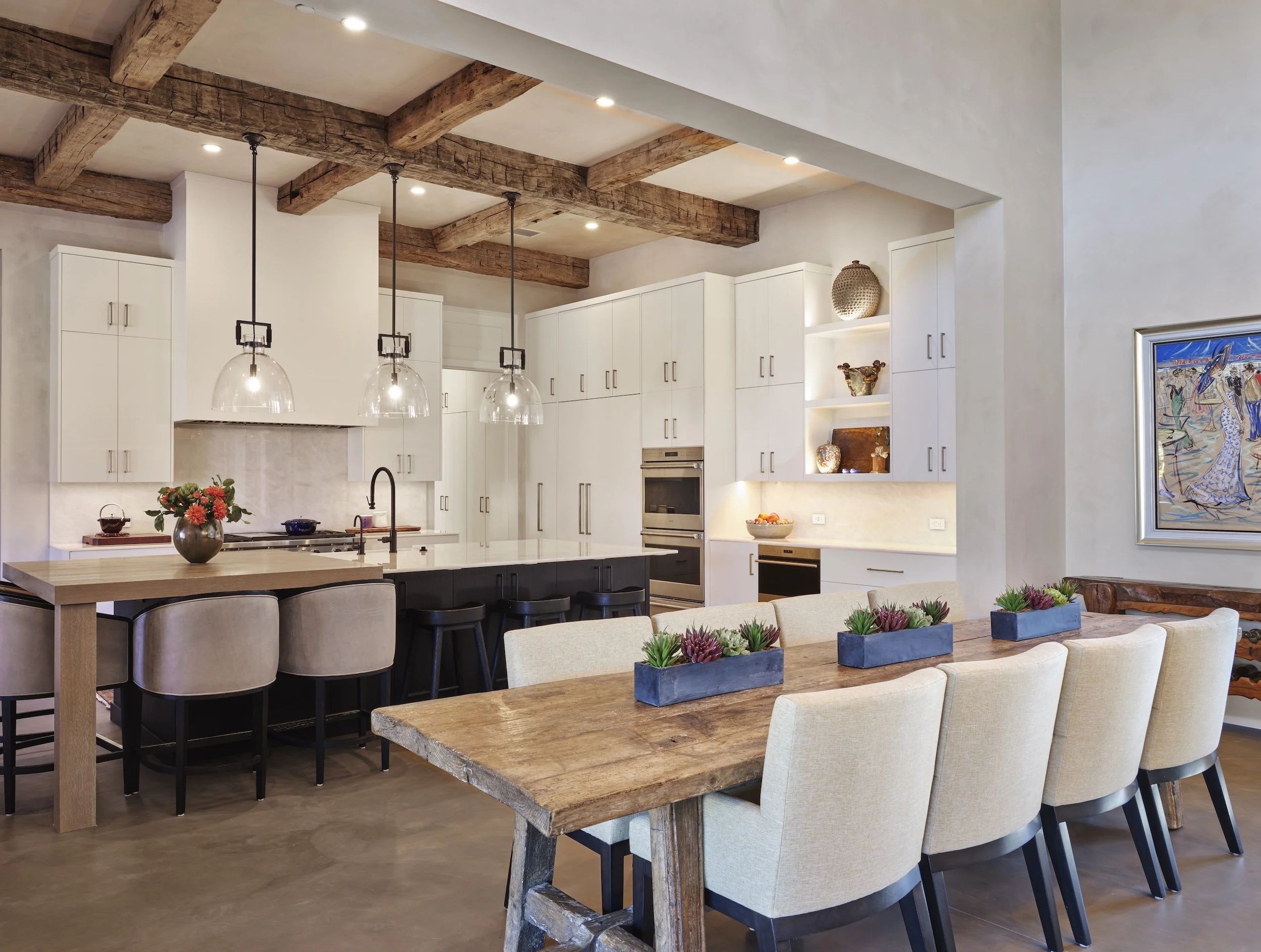Modern kitchen with white cabinets, wooden ceiling beams, black island with barstools, beige dining chairs, and a rustic wooden dining table decorated with three planters of succulents.