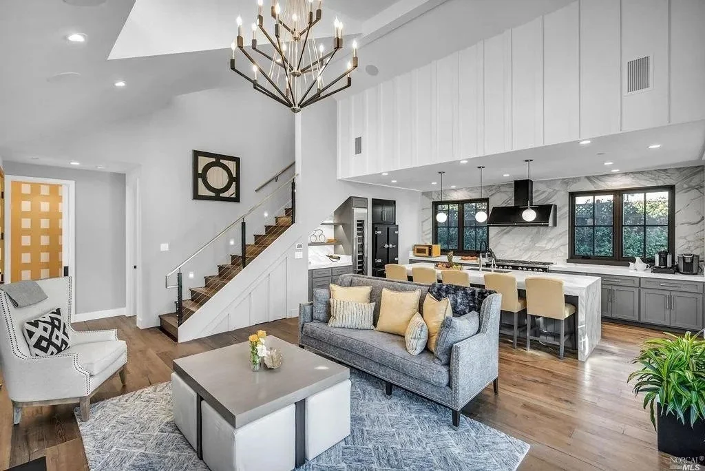 Open-concept living room and kitchen with a modern chandelier, gray and white color scheme, hardwood floors, and large windows.