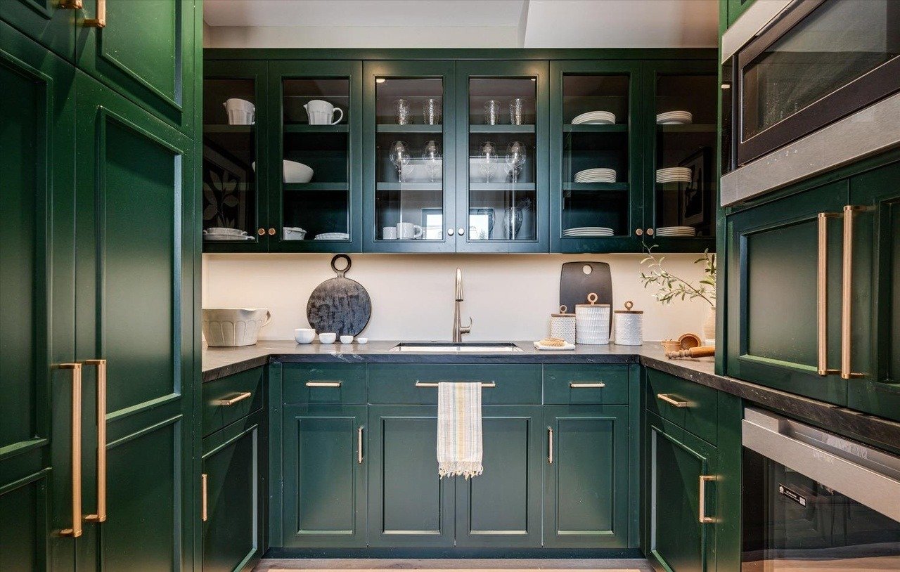 Kitchen with dark green cabinets, beige countertop, and white dishes displayed in glass cabinets above the sink. There is a towel hanging from the cabinet handle below the sink, a round cutting board, white jars, and a plant on the countertop.