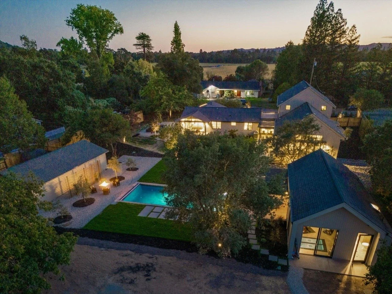 Aerial view of a residential backyard at dusk with a lit swimming pool, surrounding patio with patio furniture, and multiple houses with lit windows amid trees and greenery.
