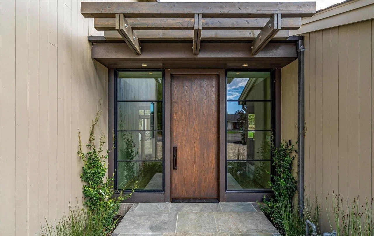 Front view of a modern house entrance with a wooden door, flanked by large glass windows, and a small garden with plants on each side.