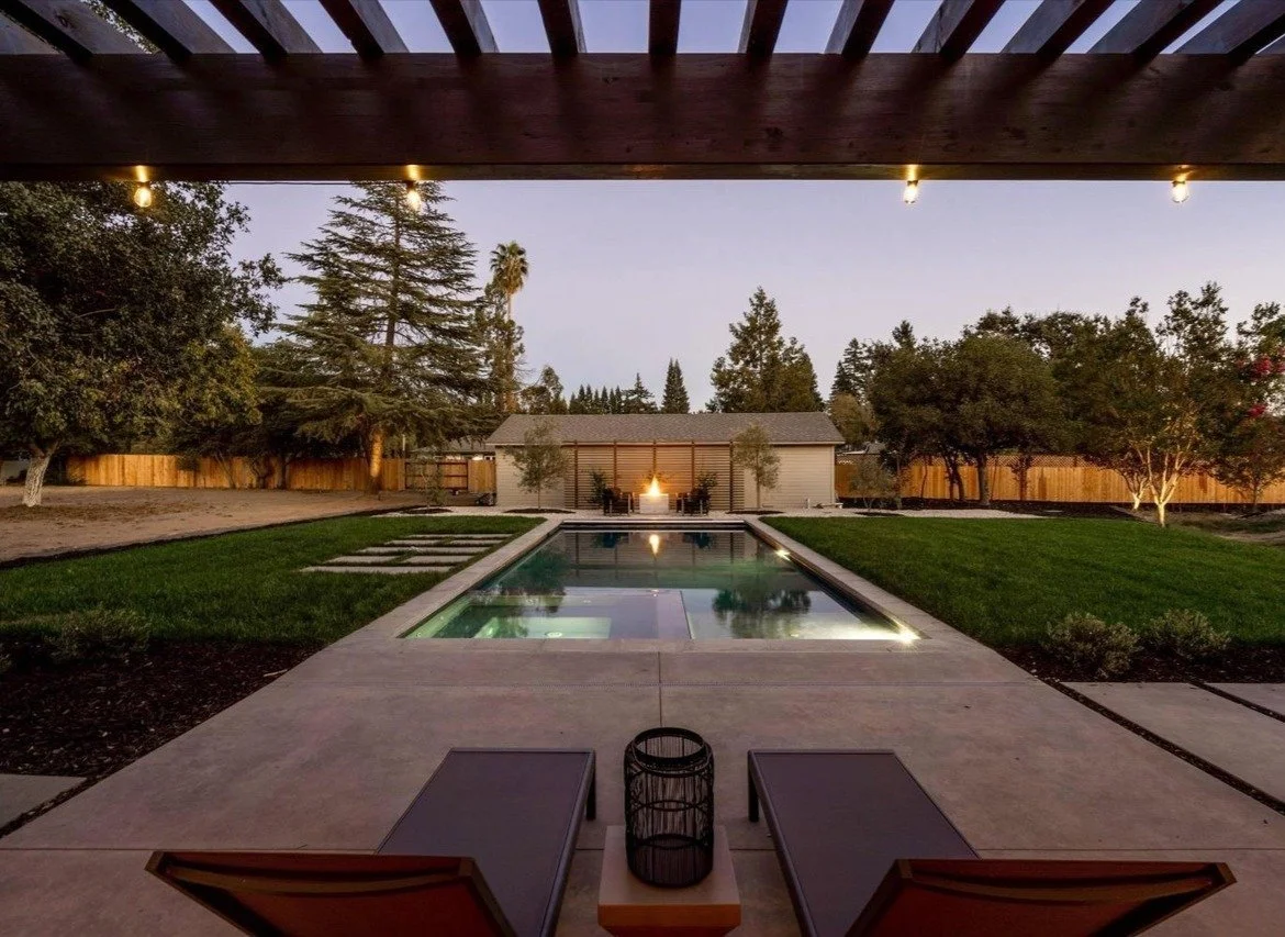 Backyard with a rectangular swimming pool, surrounded by green grass, trees, and a wooden privacy fence; outdoor seating area with two lounge chairs and a small table at the patio's edge.