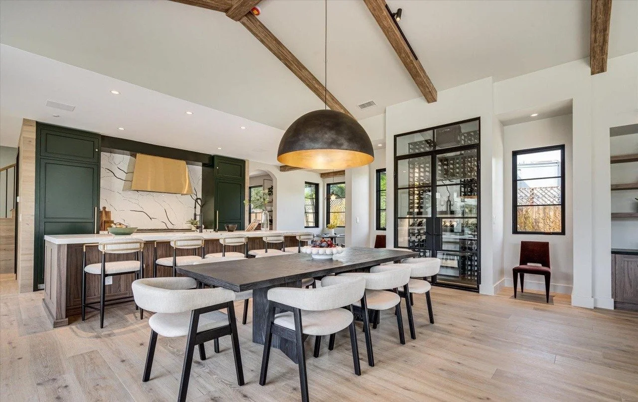 Modern kitchen and dining area with a large dining table, cream chairs, a black pendant light, green cabinets, a marble backsplash, and a wine storage glass enclosure.