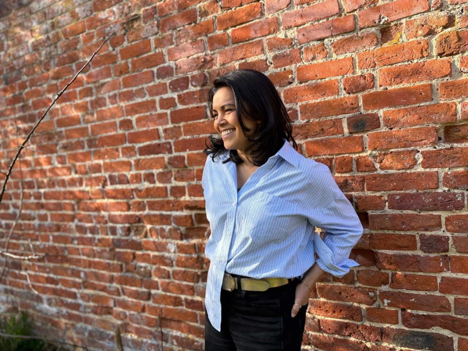 A woman with dark hair in a light blue collared shirt, smiling and leaning against a red brick wall outdoors.