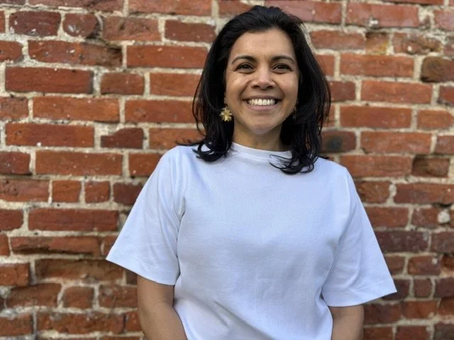 A woman with black hair wearing a white t-shirt and gold earrings, smiling in front of a red brick wall.