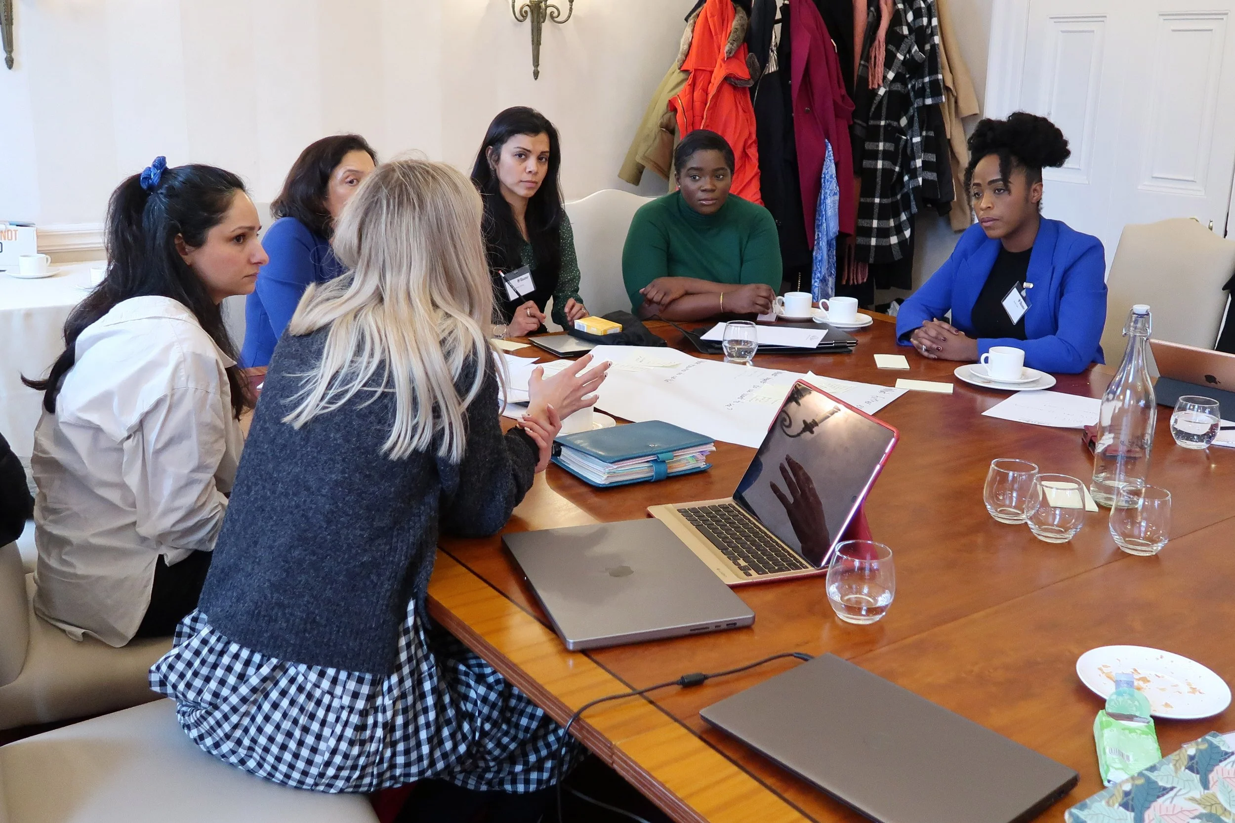 Six women sitting around a large wooden conference table having a discussion. Some women have notepads and laptops in front of them, and there are cups, glasses of water, and plates on the table. A coat rack with colorful jackets is visible in the background.