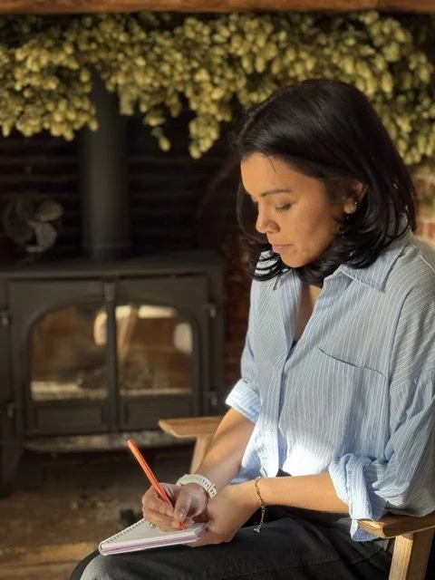 A woman with dark hair and a blue striped shirt writing in a notebook, sitting in front of a fireplace with yellow flowers above.