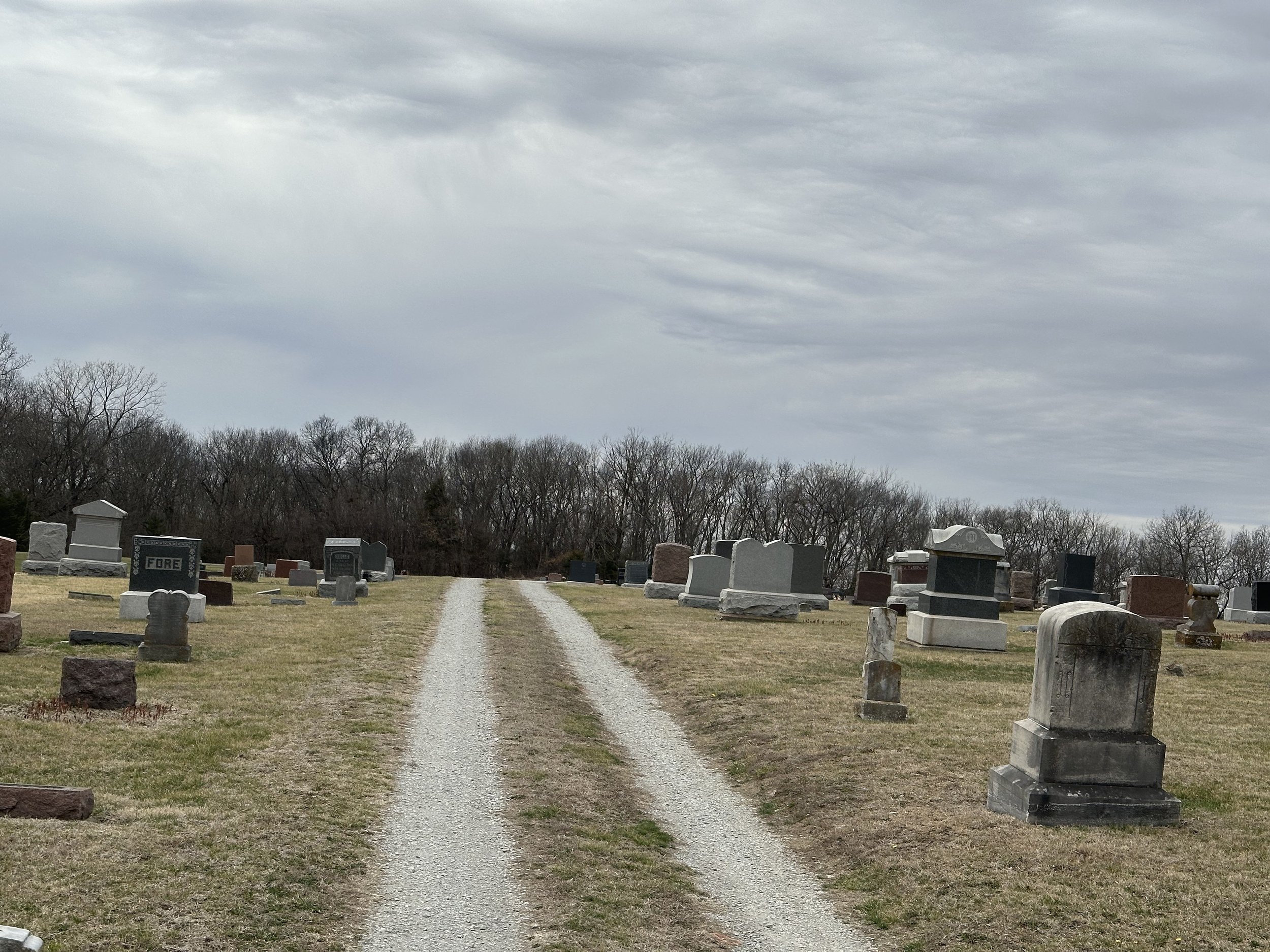 Gravel driveway in a cemetery with numerous headstones lined along both sides under an overcast sky, with leafless trees in the background.