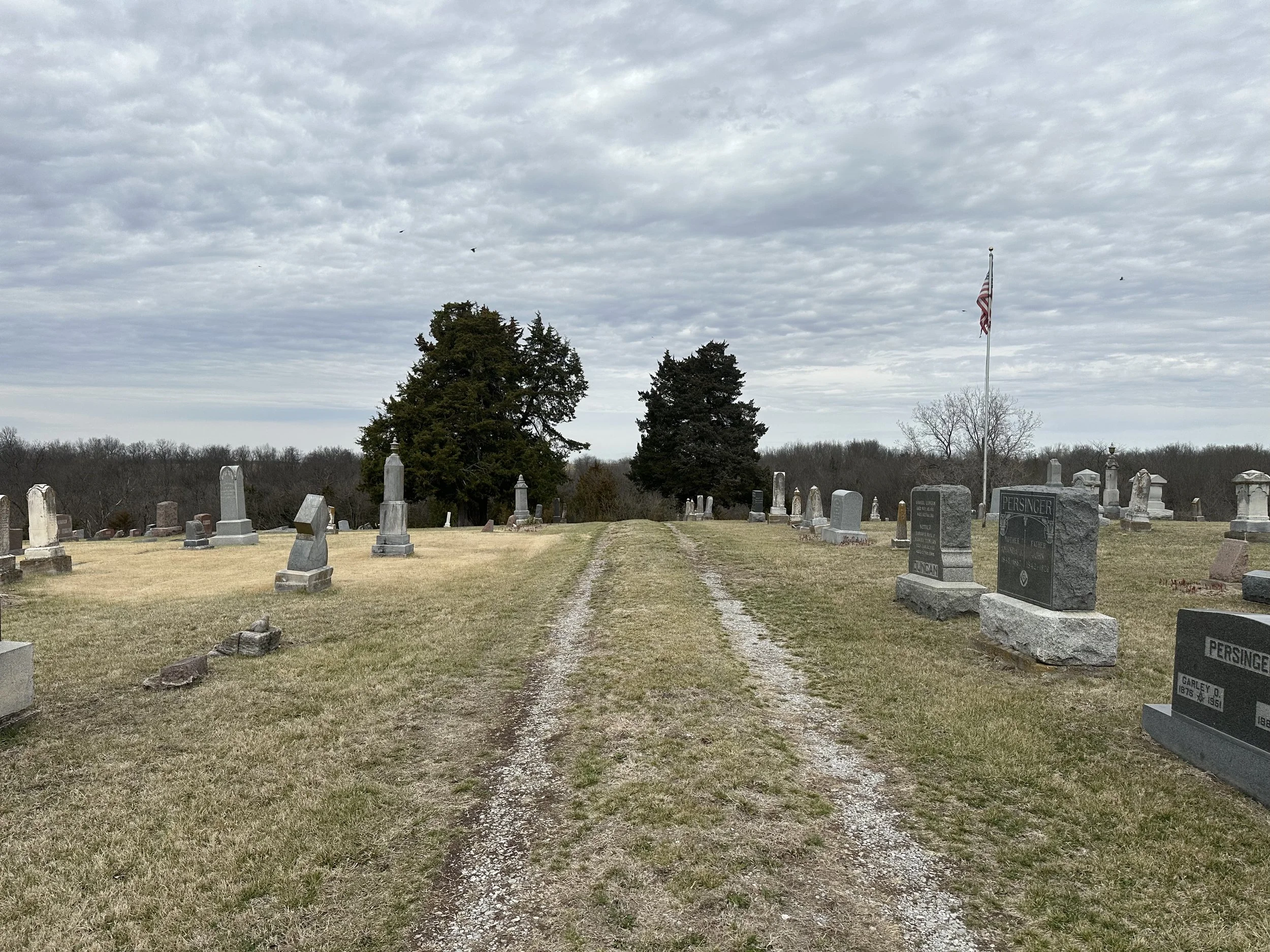 A cemetery with rows of tombstones on either side of a gravel path, under a cloudy sky, with some trees and an American flag flying in the background.