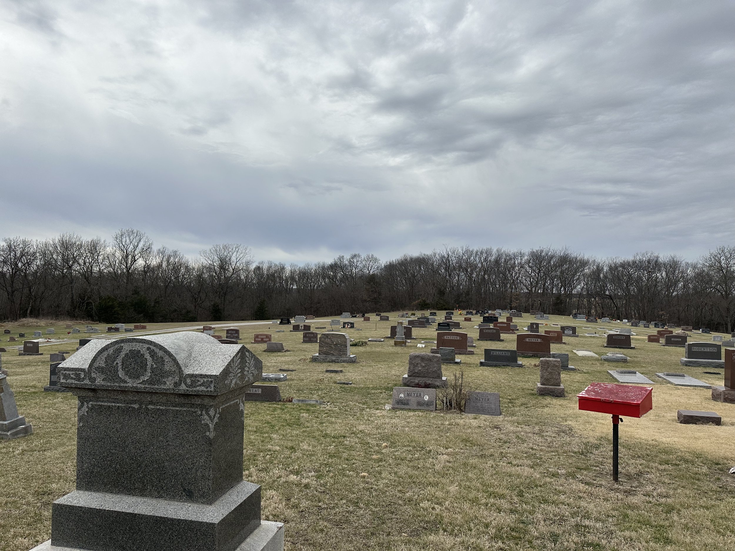 A cemetery with numerous gravestones on a grassy field under a cloudy sky, with trees in the background.