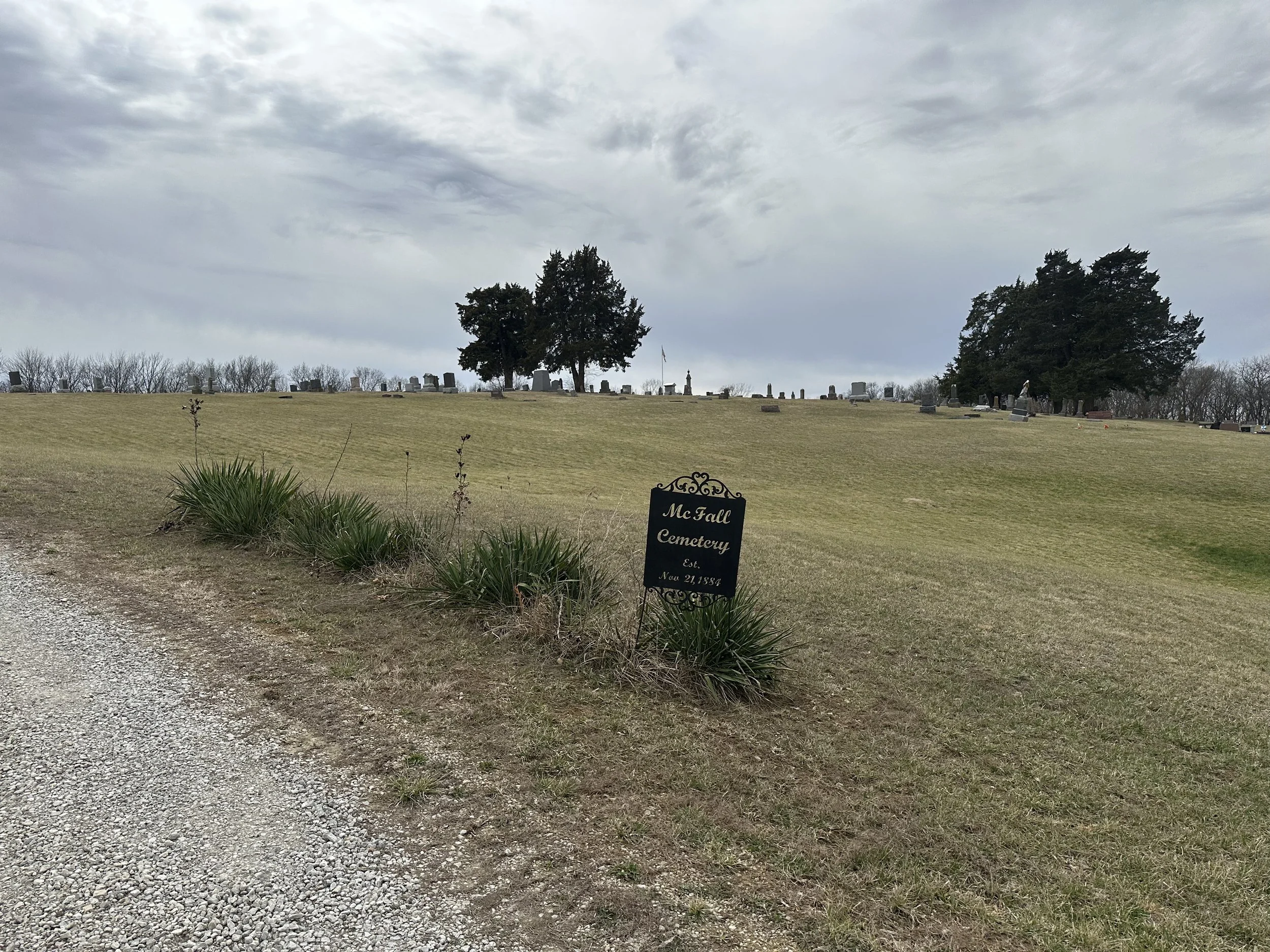 A cemetery with a sign reading 'McFall Cemetery est. Nov 21, 1884' in the foreground, grassy hill with numerous tombstones and trees in the background under a cloudy sky.